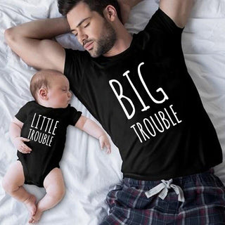 Matching father-child outfits with "Big Trouble" and "Little Trouble" text, showcasing a playful and affectionate bond between parent and baby on a cozy bed.