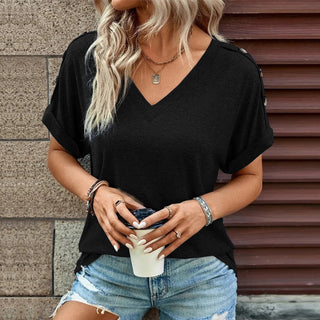 Casual woman's black V-neck top, blonde wavy hair, accessories, and jeans against a red wooden background.
