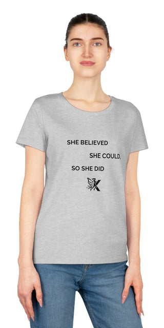 This image shows a woman wearing a gray t-shirt with text printed on it. The text reads "SHE BELIEVED SHE COULD SO SHE DID" along with a small design element. The woman in the image has a neutral facial expression and her hair is tied back. The background is plain and white.