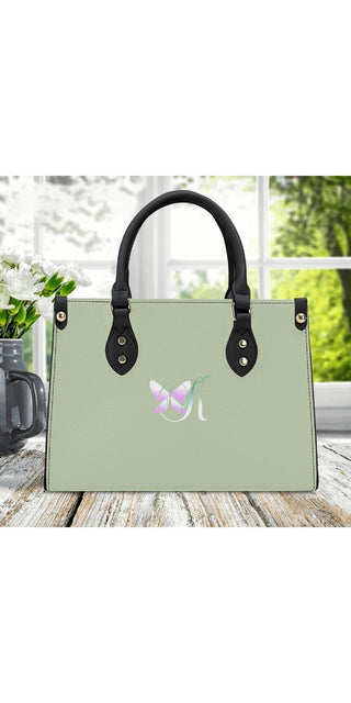 Elegant green handbag with butterfly logo, leather handles and stylish design, displayed on a wooden surface with a nature scene in the background.