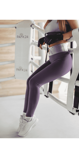 High-waist purple yoga leggings with adjustable waistband, worn by a person exercising on a gym machine with metal frame in the background.