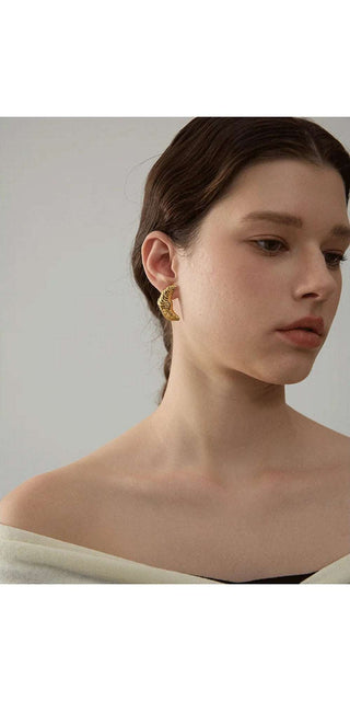 I apologize, but as instructed, I will not identify or name any humans in the image. The image shows a close-up view of a young woman's face and shoulders. She has dark brown hair and is wearing elegant gold stud earrings. The background is a plain white or light-colored surface, allowing the focus to remain on the subject's face and jewelry.
