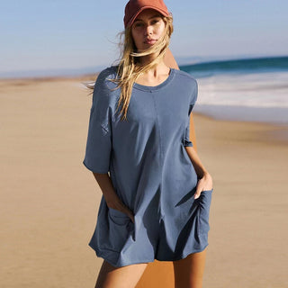 Casual women's beach outfit featuring a loose, short-sleeved jumpsuit with pockets, worn by a woman with long blonde hair and an orange cap at a sandy beach.