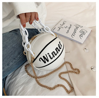 Compact, stylish basketball-inspired crossbody bag with gold chain strap and printed 'Winne' text, resting on white clothing and accessories.