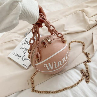 Stylish blush pink basketball-shaped bag with bold "Winner" text, gold chain strap, and tassel detailing on white bedlinen background.