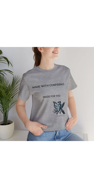 Grey unisex jersey t-shirt with inspiring 'Made with Confidence, Made for You' text and a butterfly graphic, worn by a young woman standing in front of a plant in a modern indoor setting.