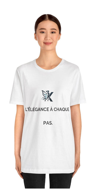 A white unisex jersey short sleeve T-shirt with the text "L'elegance a chaque pas." printed on the front. The T-shirt is worn by a smiling woman with dark hair against a plain white background.