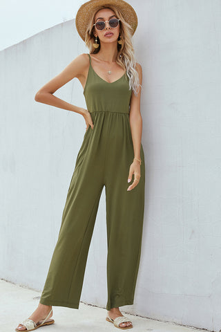 Stylish wide-leg olive jumpsuit with a sleeveless design, worn with straw hat and sunglasses against a white wall backdrop.