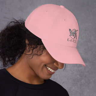 Stylish pink dad hat with K-AROLE logo on the front, worn by a smiling woman with curly dark hair against a grey background.