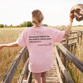 Attractive young woman wearing a pink t-shirt with an inspirational slogan printed on the back, posing on a wooden footbridge in a grassy field.