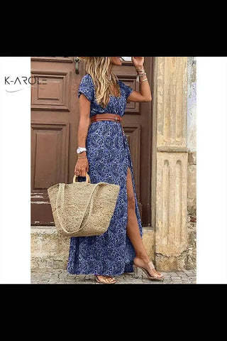 Beautiful blue printed maxi dress with high slit, worn by a female model posing in front of a wooden door backdrop, carrying a straw tote bag, showcasing K-AROLE premium fashion apparel.