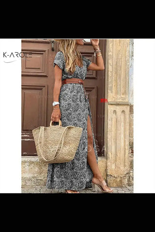 Elegant floral maxi dress with slit, straw handbag, and sandals on model posing by wooden door with K-AROLE branding displayed.