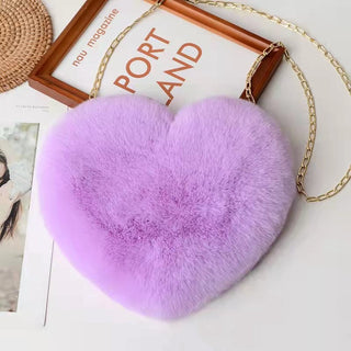 Plush heart-shaped purse in a vibrant lavender color, attached to a metallic chain strap, displayed on a wooden plaque with text against a white background.