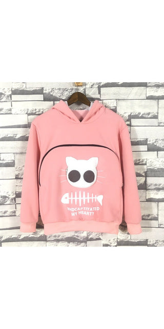 Cozy pink hooded sweatshirt with white cat skull graphic on front, hanging on a wooden hanger against a brick wall background.