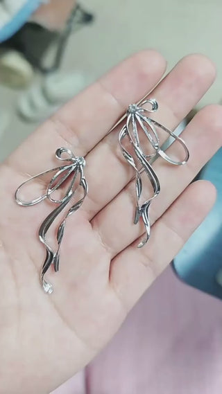 Elegant silver-toned irregular large bow earrings with tassel streamers, displayed in a person's hand against a soft background.