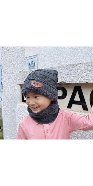 Young girl wearing a stylish knitted winter hat and scarf, standing in front of a concrete wall with text on it.