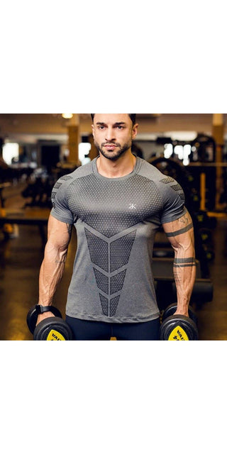 Muscular male model wearing form-fitting grey athletic compression t-shirt with geometric pattern, showcasing his fit physique in a gym setting.