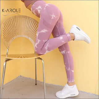 Stylish woman wearing pink tie-dye patterned yoga tights and white sneakers, posing on a modern chair in front of the K-AROLE brand logo.
