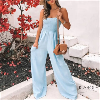 Stylish women's casual light blue jumpsuit with a smocked bodice, displayed against a backdrop of vibrant red flowers on a branch. The jumpsuit features a sleeveless design and a flowing, wide-leg silhouette, creating a chic and comfortable look.